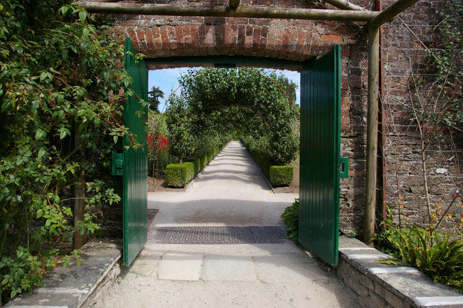 A winding garden pathway paved with light-colored bricks, bordered by neatly trimmed green hedges on both sides. Overhanging deciduous trees display autumn foliage with leaves in shades of green, yell
