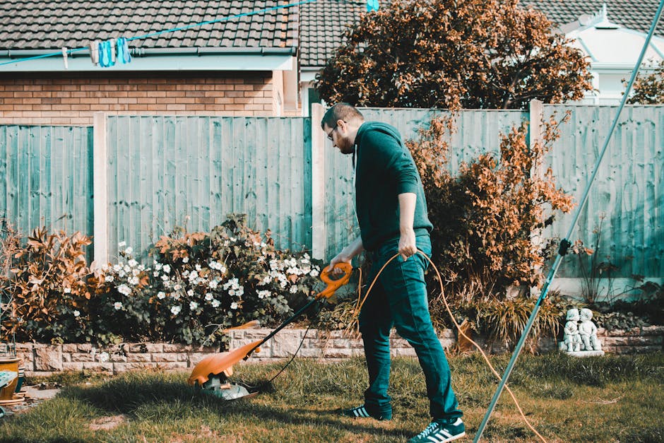 A man trimming grass in a small backyard garden during daytime. He wears glasses, a dark hoodie, and jeans while operating a cordless grass trimmer with an orange body and black handle, which is conne